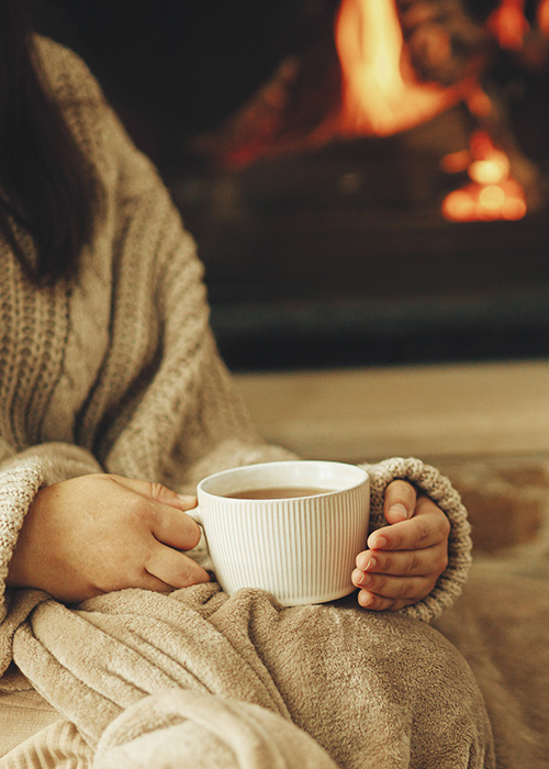 Frau mit Decke und Heißgetränk vor brennendem Kamin